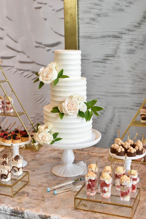 Three-tier textured white wedding cake with blush roses displayed on dessert table with treats and gold accents