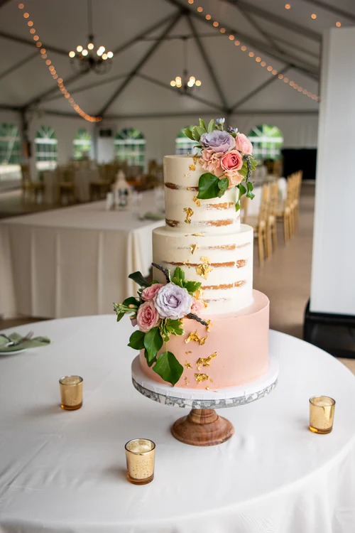 Three-tier wedding cake with semi-naked frosting, blush pink bottom tier, and purple and pink roses in tented reception venue