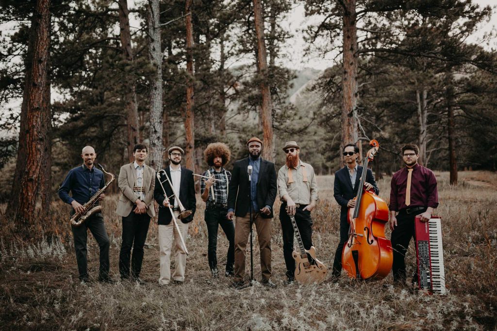 Eight musicians with saxophones, guitars, upright bass, and keyboard standing in pine forest clearing