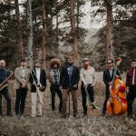 Eight musicians with saxophones, guitars, upright bass, and keyboard standing in pine forest clearing