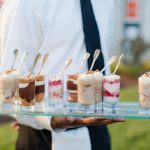 Server holding tray of layered dessert shots in small glasses with spoons at outdoor event