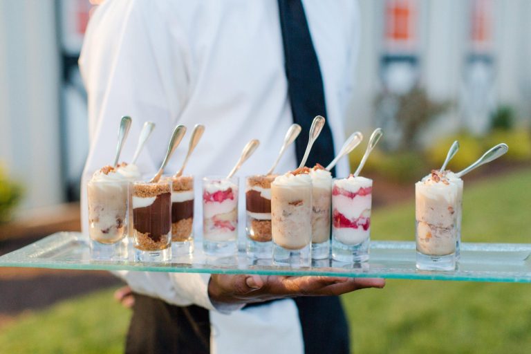 Server holding tray of layered dessert shots in small glasses with spoons at outdoor event