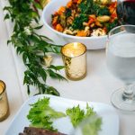 Elegant catered table setting with colorful vegetable bowl, fresh salad, wine glasses, and gold votives with greenery