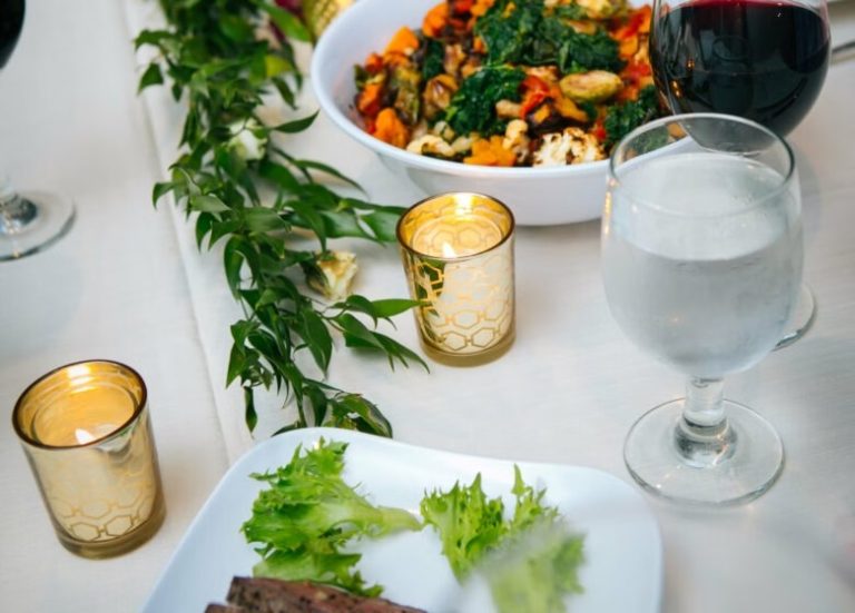 Elegant catered table setting with colorful vegetable bowl, fresh salad, wine glasses, and gold votives with greenery