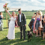 Newlyweds walking down aisle with black dog while guests blow bubbles at outdoor wedding celebration