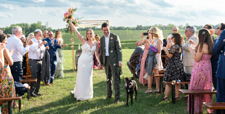 Newlyweds walking down aisle with black dog while guests blow bubbles at outdoor wedding celebration