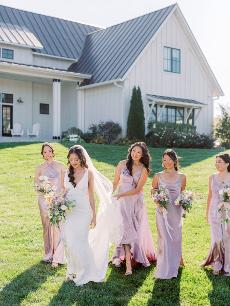 Bridal party in mauve dresses walking together on lawn in front of contemporary white barn venue