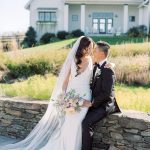 Married couple sitting intimately on stacked stone wall with modern white farmhouse and natural prairie grasses in background