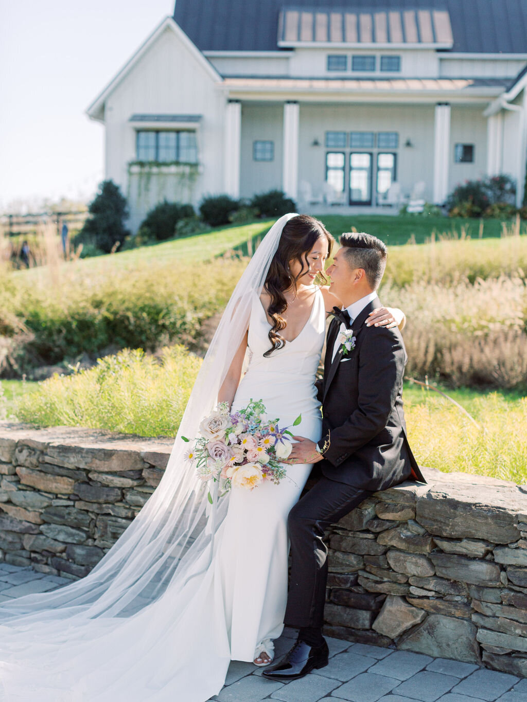 Married couple sitting intimately on stacked stone wall with modern white farmhouse and natural prairie grasses in background
