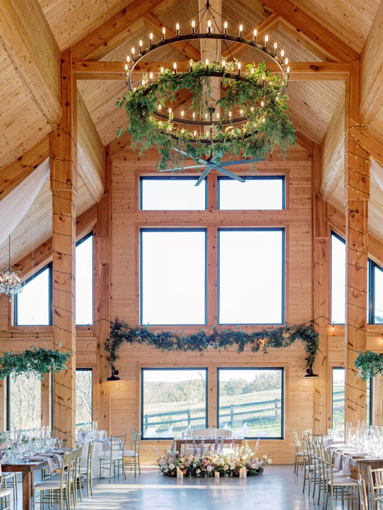 Cathedral ceiling barn reception hall with candle chandelier, greenery garland, and pastoral countryside views through floor-to-ceiling windows