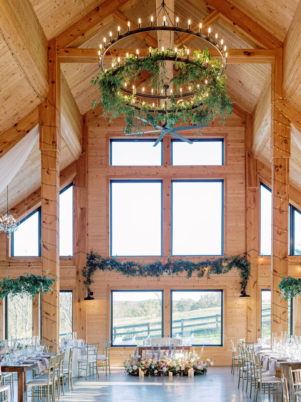 Cathedral ceiling barn reception hall with candle chandelier, greenery garland, and pastoral countryside views through floor-to-ceiling windows