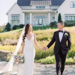 Bride and groom holding hands walking toward modern farmhouse with white siding and landscaped grounds