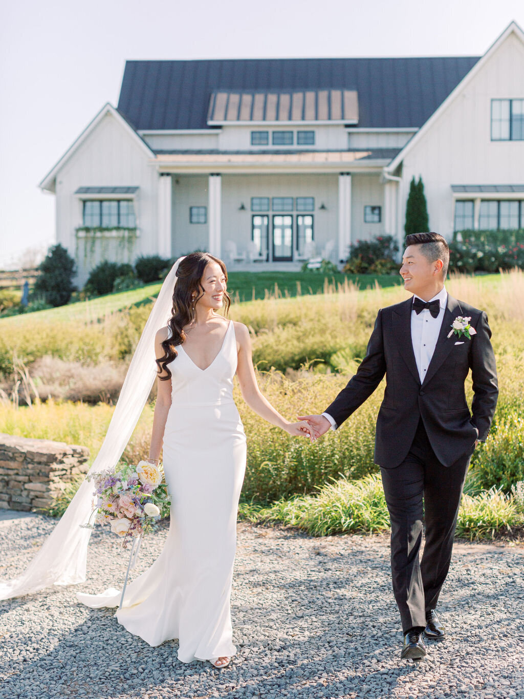 Bride and groom holding hands walking toward modern farmhouse with white siding and landscaped grounds