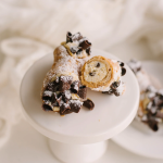 Chocolate chip cannoli dusted with powdered sugar on white cake stand