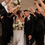 Smiling newlyweds exit under arch of white bouquets held by wedding party highlighting bride's elegant updo and makeup