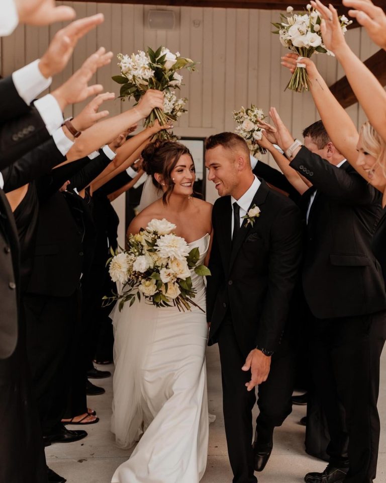 Smiling newlyweds exit under arch of white bouquets held by wedding party highlighting bride's elegant updo and makeup