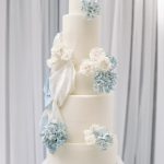 Five-tier white wedding cake adorned with ivory roses and dusty blue hydrangeas cascading diagonally down the tiers
