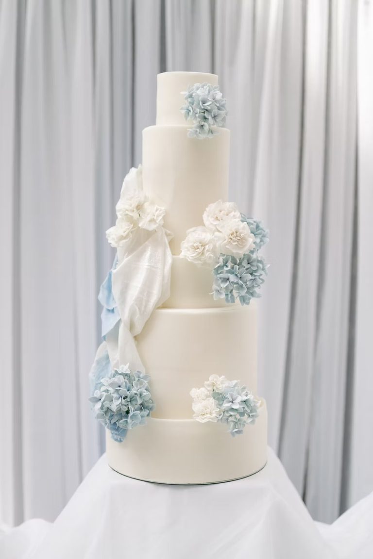 Five-tier white wedding cake adorned with ivory roses and dusty blue hydrangeas cascading diagonally down the tiers