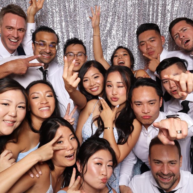 Wedding guests making peace signs and silly faces in photo booth with silver sequin backdrop