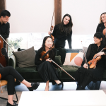 String ensemble of six musicians in black attire with violins and cellos gathered in modern lounge setting