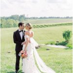 Newlyweds standing close together on grassy hill with vineyard and pond in background