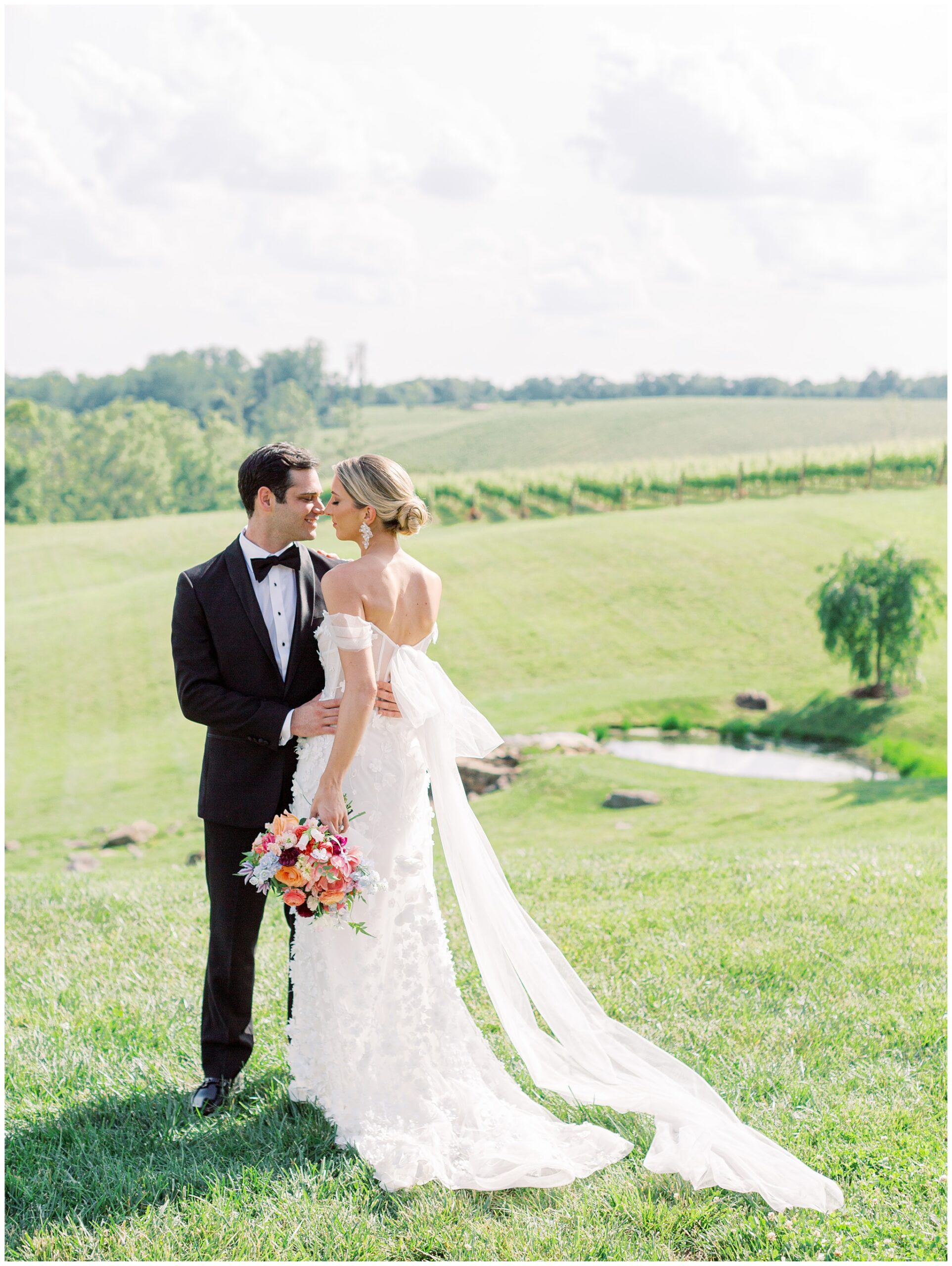 Newlyweds standing close together on grassy hill with vineyard and pond in background