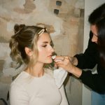 Makeup artist applying lipstick to bride with styled updo wearing cream sweater