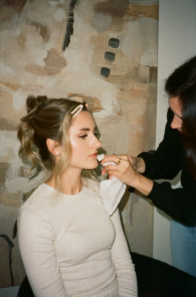 Makeup artist applying lipstick to bride with styled updo wearing cream sweater