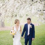 Bride in v-neck gown and groom in navy suit beneath flowering trees
