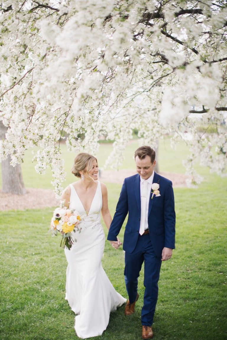 Bride in v-neck gown and groom in navy suit beneath flowering trees