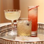 Three craft cocktails on decorative brass tray including coupe glass, rocks glass, and highball with citrus garnish