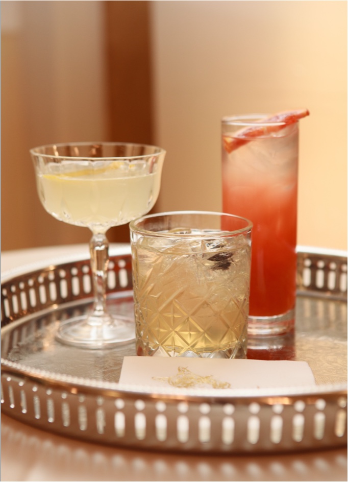 Three craft cocktails on decorative brass tray including coupe glass, rocks glass, and highball with citrus garnish