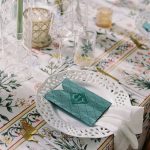 Elegant table setting with white scalloped plate, teal quilted favor envelope, botanical printed tablecloth, and assorted glassware