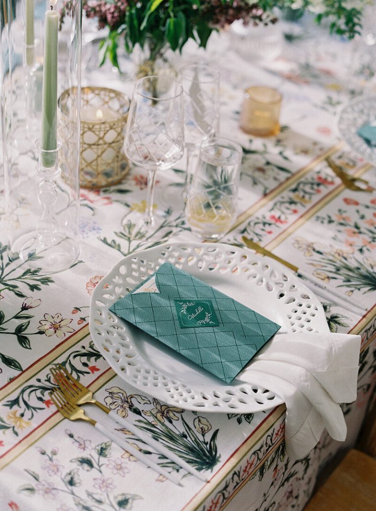 Elegant table setting with white scalloped plate, teal quilted favor envelope, botanical printed tablecloth, and assorted glassware