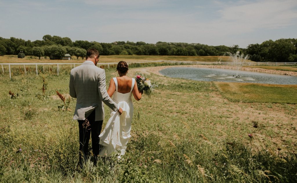 Bride and groom walking hand-in-hand through Nebraska countryside with pond and trees in background