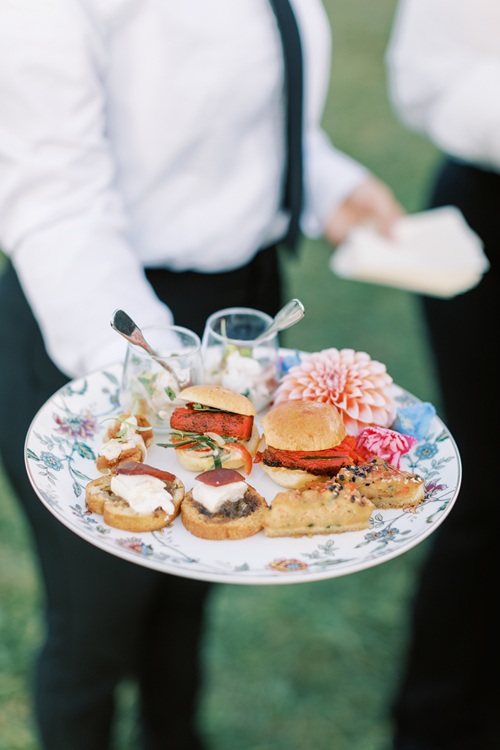 Server holding floral china plate with assorted appetizers including sliders and crostini at wedding reception