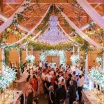 Lavishly decorated barn venue with exposed beams, chandelier, greenery, and illuminated floral archway