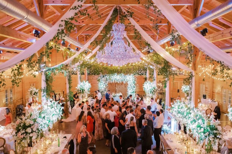 Lavishly decorated barn venue with exposed beams, chandelier, greenery, and illuminated floral archway