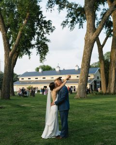 Bride and groom embracing on lawn under mature trees with barn-style venue and guests in background