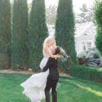 Groom lifts bride in flowing white dress for romantic kiss among tall evergreen trees and manicured garden