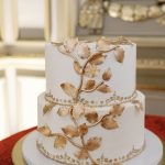 Two-tier white wedding cake adorned with gold leaf branches and pearl border details on gold cake stand
