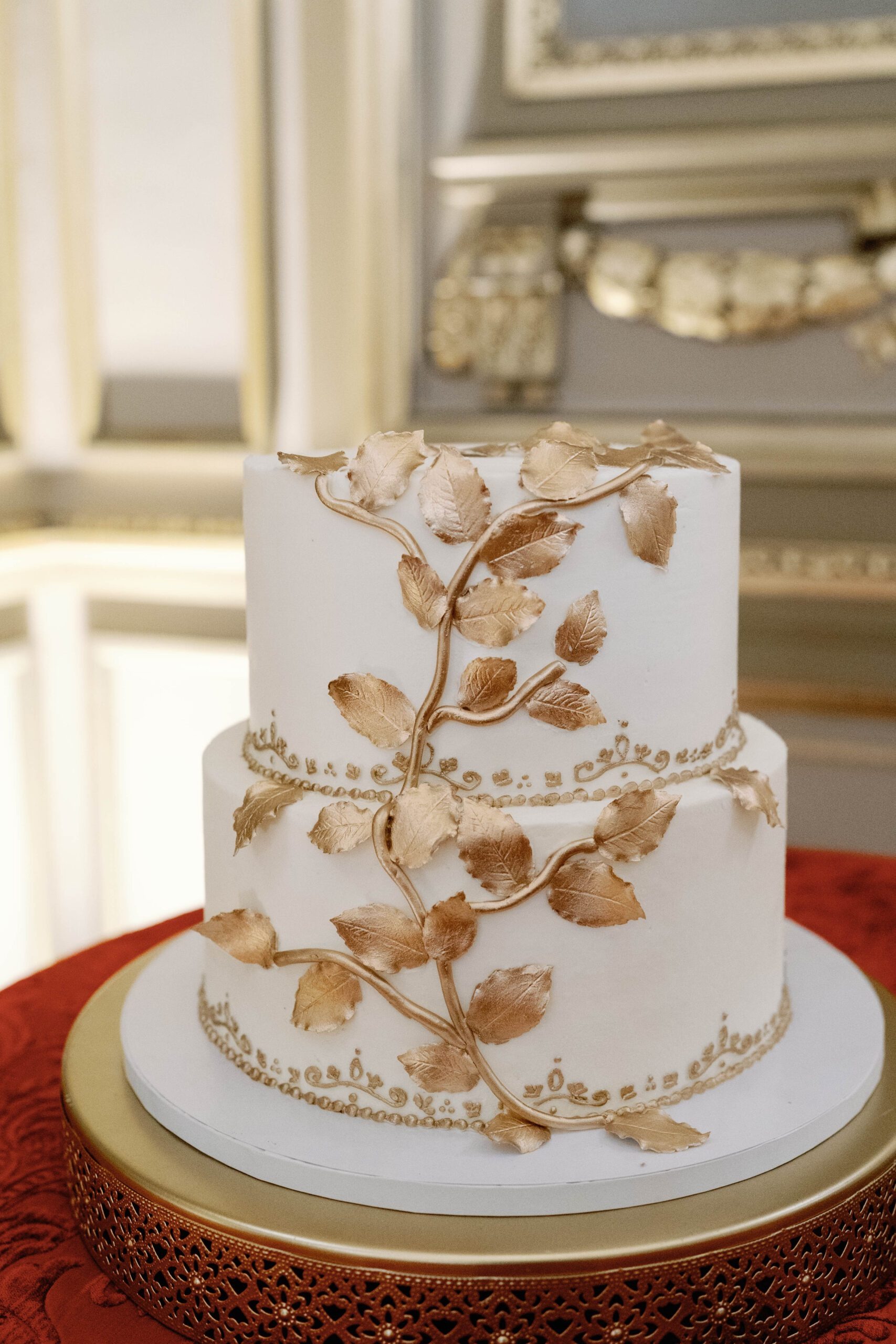 Two-tier white wedding cake adorned with gold leaf branches and pearl border details on gold cake stand