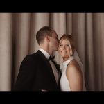 Groom kissing bride's temple as she smiles at camera in elegant indoor setting