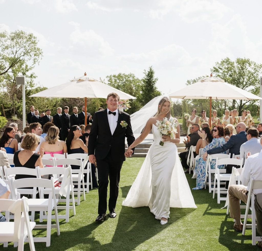 Newlyweds walking down outdoor ceremony aisle under white umbrellas with wedding party and guests applauding