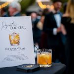 Signature cocktail card beside glass of whiskey at formal event with blurred guests in background