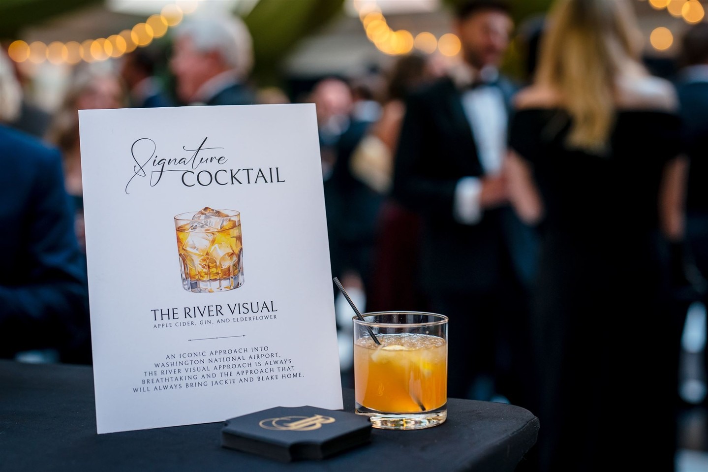 Signature cocktail card beside glass of whiskey at formal event with blurred guests in background
