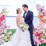 Newlyweds standing between elaborate floral pillars in pink, coral, and white with waterfront backdrop