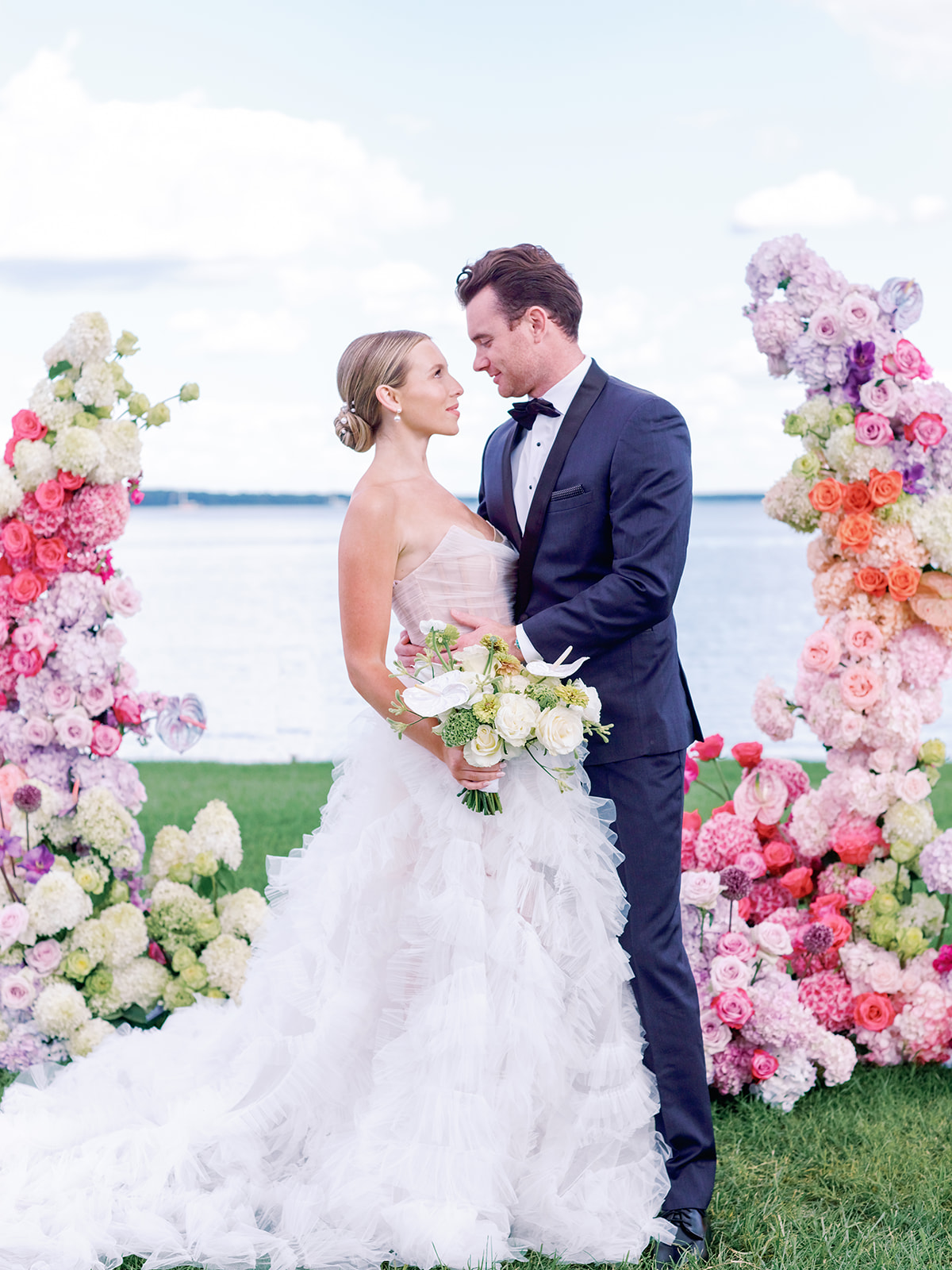 Newlyweds standing between elaborate floral pillars in pink, coral, and white with waterfront backdrop