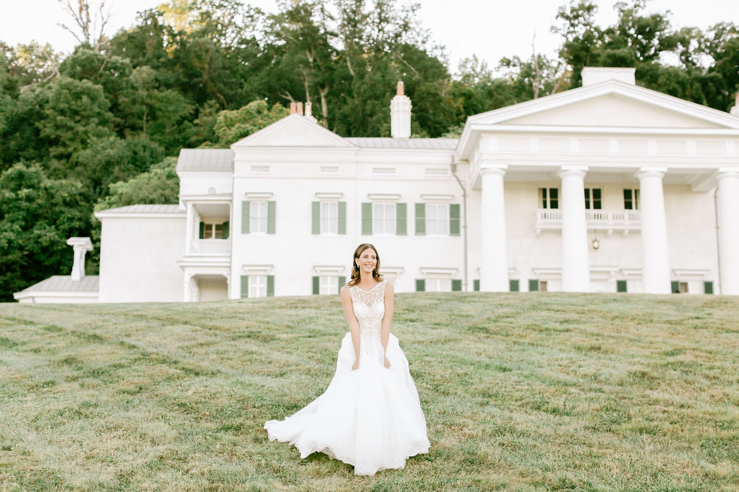 Bride in flowing white ball gown standing on lawn in front of white colonial-style estate venue
