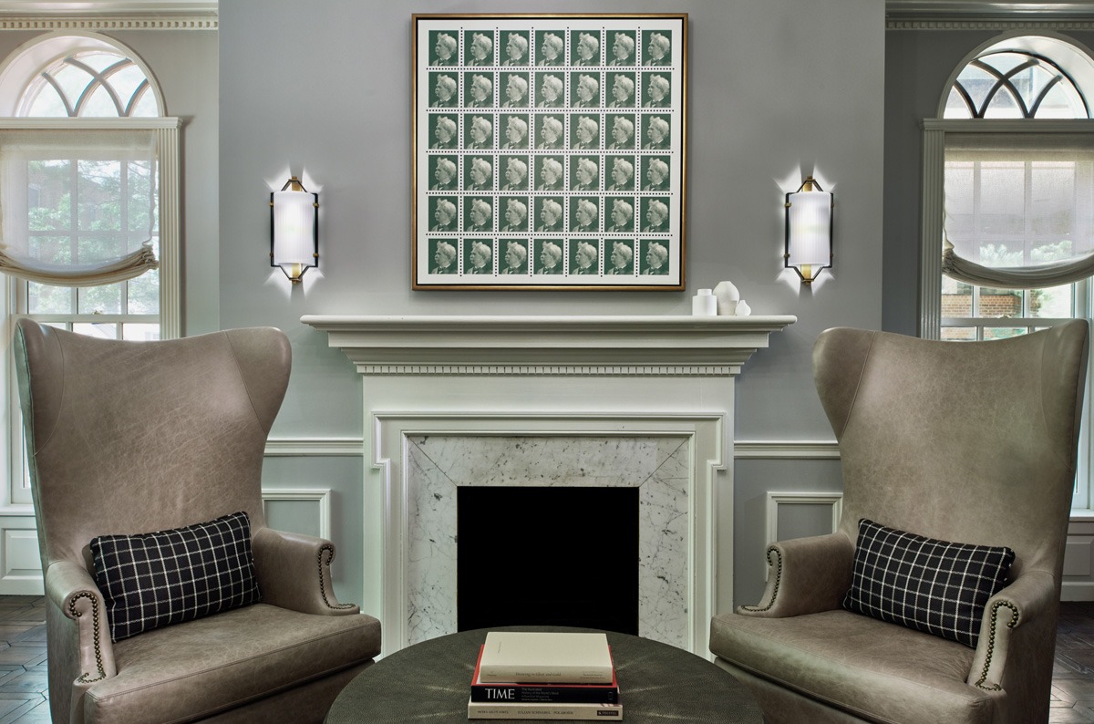 Symmetrical seating area with taupe wingback chairs, framed stamp collection above marble fireplace mantel